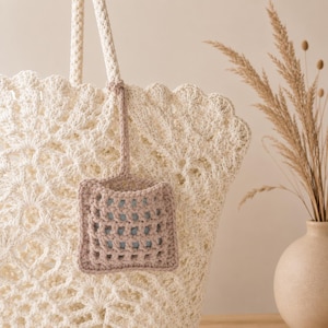 May include: A cream-colored crocheted tote bag with a scalloped top edge and thick braided handles. A small, square, light brown crocheted pouch with an open grid pattern hangs from one of the handles, revealing a pale blue interior lining.