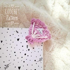 May include: A handmade, crochet bookmark in a soft pink hue, adorned with white stitching and a small, decorative square. The bookmark is placed on a book with a black and white dotted pattern. The text "CROCHET Pattern" is visible.