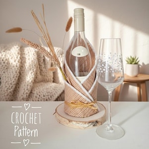 May include: A clear wine bottle is encased in a beige and white crocheted cozy, tied with raffia, and set on a wooden slice. A wine glass with a dotted design stands beside it. The words "CROCHET Pattern" are written in white.