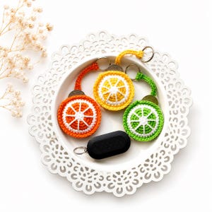 Puede incluir: Tres llaveros circulares de ganchillo con forma de rodajas de cítricos en naranja, amarillo y verde, cada uno con un centro blanco y una anilla de metal. Un llavero ovalado de color negro sólido con una anilla de metal se sitúa debajo.