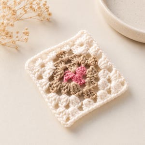 May include: A square crochet granny square motif featuring a central pink cluster, surrounded by a layer of light brown yarn, and finished with an outer border of cream-coloured yarn. The piece displays a classic openwork stitch pattern.