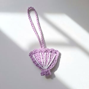 May include: A handmade, crochet lavender fan-shaped ornament with white accents. The ornament has a looped lavender cord for hanging. The fan design features a scalloped edge and white vertical lines, creating a textured, decorative piece.