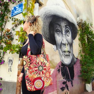 May include: A woman in a black shirt and a colorful tote bag stands in front of a mural of a woman wearing a hat. The mural is on a building with a street sign that reads "Rue de Lorraine".