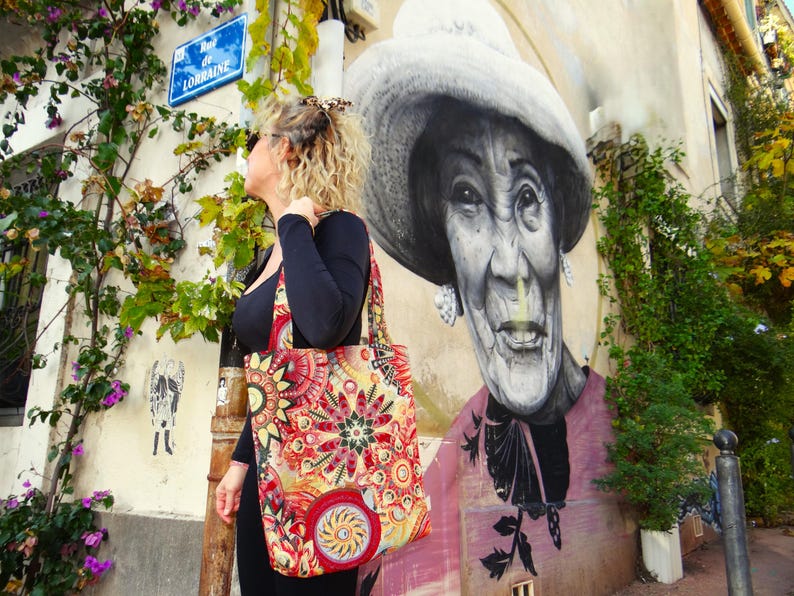 May include: A woman in a black shirt and a colorful tote bag stands in front of a mural of a woman wearing a hat. The mural is on a building with a street sign that reads "Rue de Lorraine".