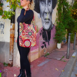 May include: A woman wearing black leggings and black boots is standing in front of a mural. She is carrying a colorful tote bag with a floral pattern. The mural depicts a woman with a hat and a serious expression.