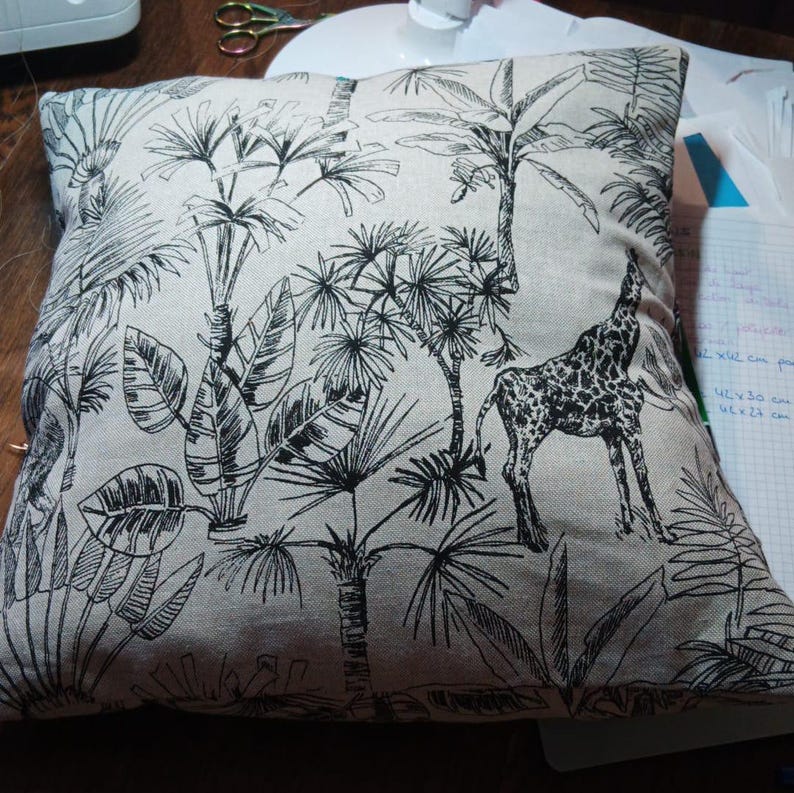 May include: A beige throw pillow with a black and white print of palm trees and a giraffe. The pillow is made of a soft, linen-like fabric.