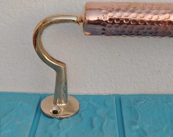 May include: A copper and brass towel bar. The bar has a hammered copper finish on the horizontal section and a curved brass support. The base is round and brass. The bar is mounted on a blue surface and a white wall.