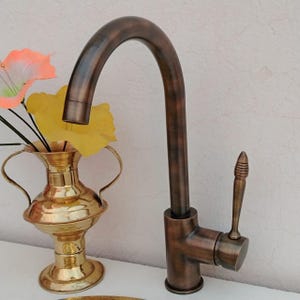 Op de afbeelding: Een bronzen keukenkraan met een gebogen uitloop en een hendel. De kraan staat tegen een witte achtergrond, naast een gouden vaas met bloemen.