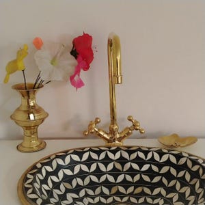 May include: A gold faucet with a black and white bone inlay oval sink. The sink has a leaf pattern. A gold vase with artificial flowers sits on the counter.