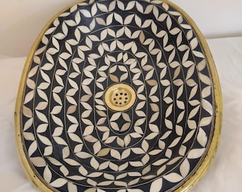 Op de afbeelding: Ovaalvormige wastafel gemaakt van zwart en wit botinleg met een messing rand. De wastafel heeft een geometrisch patroon van bladeren en een messing afvoer.