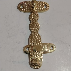 May include: A gold-colored, decorative cabinet latch with a textured, hammered finish. The latch has an elongated, vertical design with rounded ends and a central, curved section. It features a knob at each end and a pivoting arm.