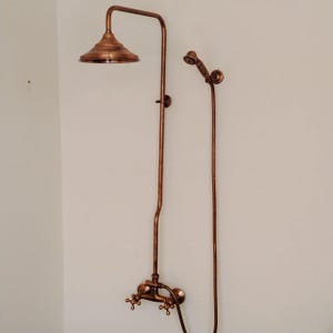 Puede incluir: Un conjunto de ducha de color cobre, que incluye un cabezal de ducha de lluvia y una ducha de mano. El accesorio está montado en la pared y presenta un diseño vintage con manijas en forma de cruz.