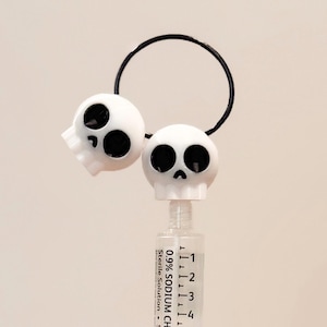 May include: Two white skull shaped bottle stoppers with black eyes and a black mouth. The stoppers are connected by a black ring. The stoppers are on top of a clear glass bottle with black markings that read "0.9% SODIUM CHLORIDE Sterile Solution".