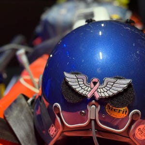 May include: A blue helmet with a silver wing patch and a pink ribbon. The helmet has a Merit sticker with a phone number. The helmet is sitting on an orange surface.