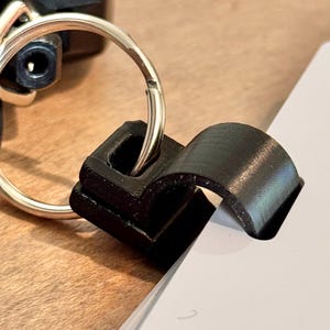 May include: A black 3D-printed keychain clip, attached to a silver key ring, is holding a stack of white paper. The clip has a curved design and is made of a matte black material. The background is a wooden surface.