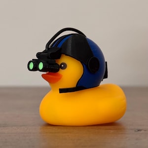 May include: A yellow rubber duck wearing a blue helmet with a black strap and green night vision goggles.
