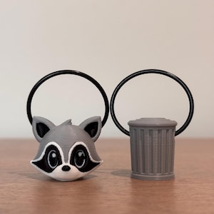 May include: Two gray 3D printed keychains, one shaped like a raccoon face and the other shaped like a trash can.