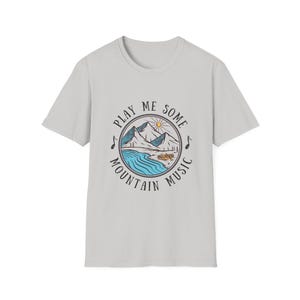 Puede incluir: Una camiseta gris claro con un estampado gráfico de una escena de montaña y el texto "Play Me Some Mountain Music".