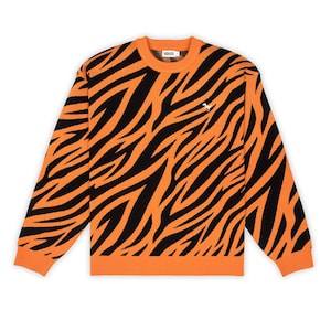 May include: A long-sleeve crewneck jumper featuring a bold orange and black tiger stripe pattern. The garment has a ribbed orange collar, cuffs, and hem. A small white embroidered dinosaur logo is positioned on the upper left chest area.
