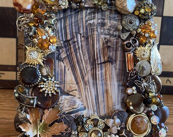 May include: A picture frame decorated with a variety of vintage jewelry, including rhinestones, buttons, and brooches. The frame is made of wood and has a brown and gold finish.