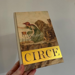 May include: A book titled "CIRCE" with a gold foil cover. The cover features an illustration of a woman in a green dress standing on a cliff with two lions.
