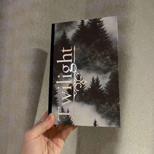 Peut inclure: Un livre reli&eacute; noir avec un motif argent&eacute; sur la couverture. Le motif repr&eacute;sente une sc&egrave;ne de for&ecirc;t avec des arbres et du brouillard, et le mot "Twilight" dans une police de caract&egrave;res stylis&eacute;e.