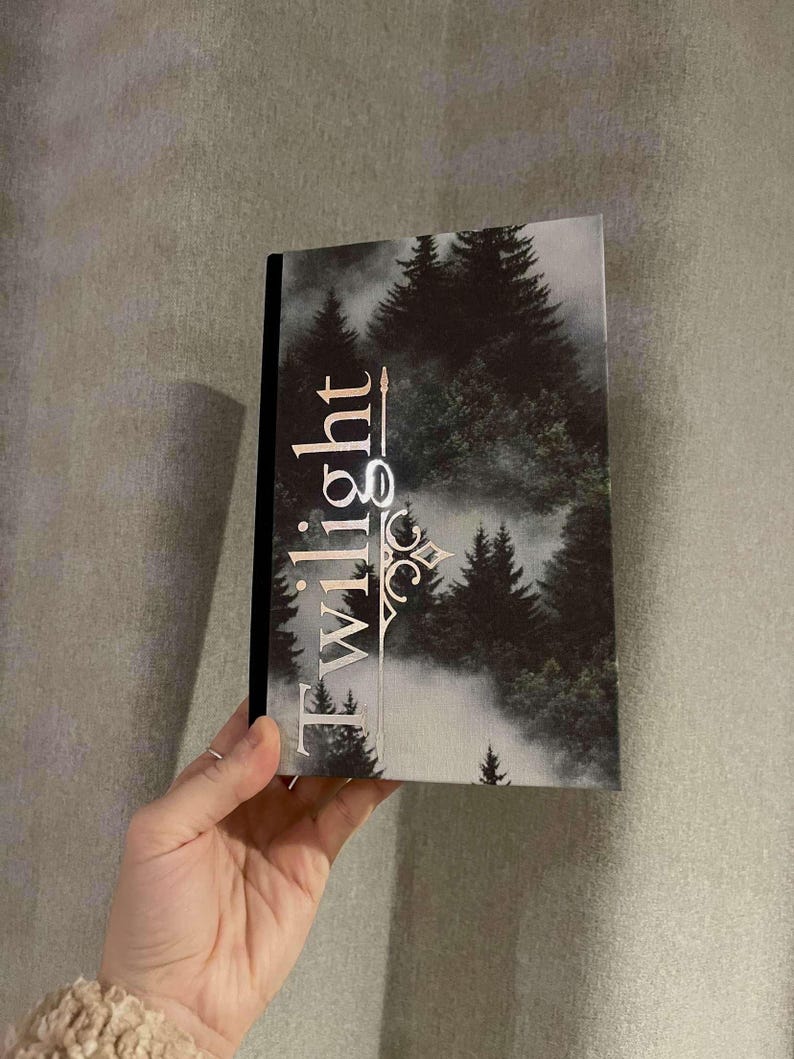 Peut inclure: Un livre reli&eacute; noir avec un motif argent&eacute; sur la couverture. Le motif repr&eacute;sente une sc&egrave;ne de for&ecirc;t avec des arbres et du brouillard, et le mot "Twilight" dans une police de caract&egrave;res stylis&eacute;e.