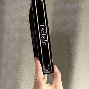 Peut inclure: Un livre reli&eacute; noir avec le titre "Twilight" imprim&eacute; en blanc sur le dos. Le livre est tenu dans une main avec une bague en argent.
