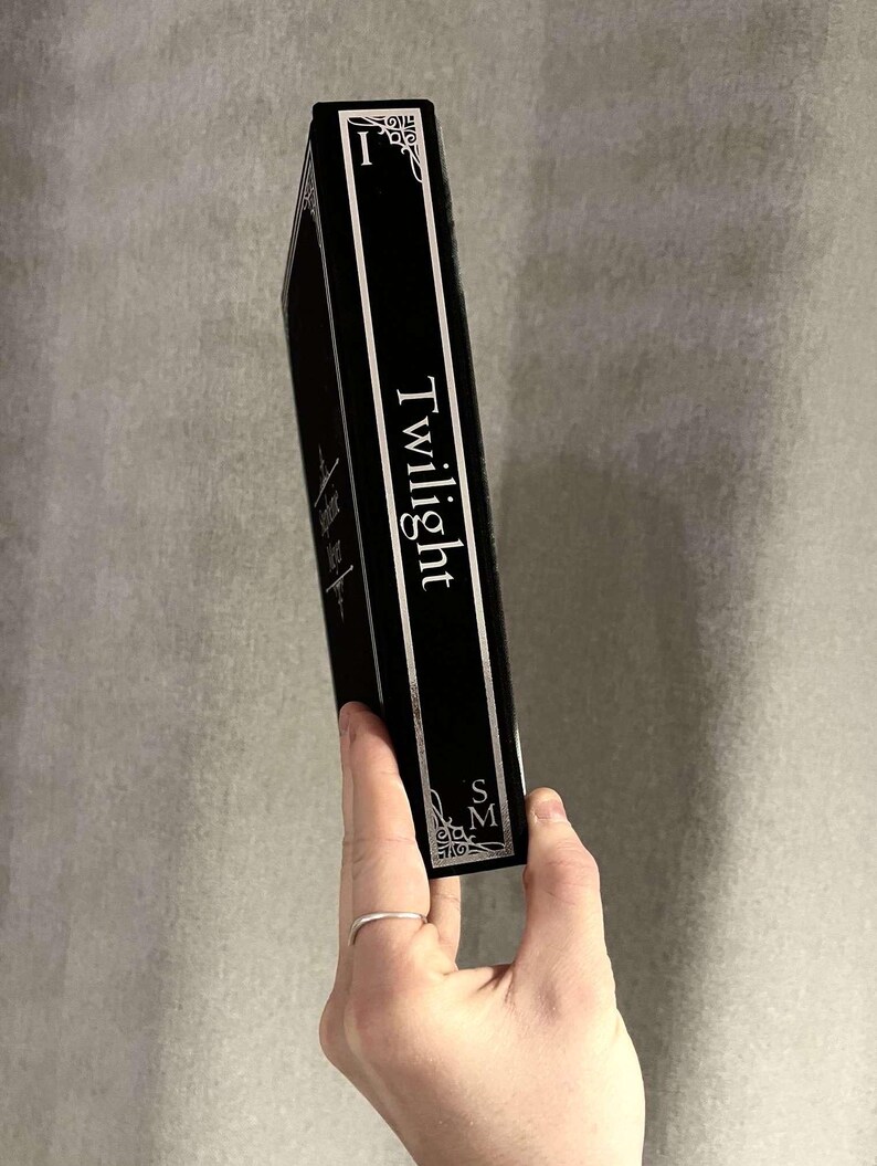 Peut inclure: Un livre reli&eacute; noir avec le titre "Twilight" imprim&eacute; en blanc sur le dos. Le livre est tenu dans une main avec une bague en argent.