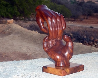 May include: A wooden sculpture of two hands clasped together, the top hand is reaching down to grasp the bottom hand. The sculpture is made of a dark reddish-brown wood and is sitting on a wooden base.