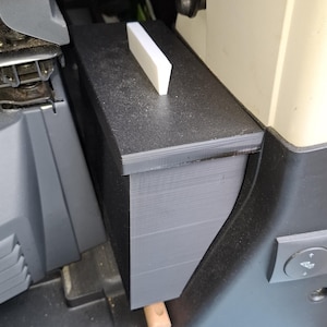 May include: A black rectangular storage container with a flat top surface and a small white vertical tab protruding from the center. The container features a smooth, matte finish and is positioned vertically within a narrow interior space.