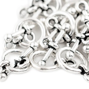 May include: A close-up of a pile of silver-toned metal toggle clasps. The clasps are designed with a round loop and a bar with a ball on each end.