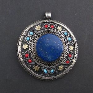 May include: A round, silver-toned pendant with a large, deep blue stone center. The pendant is adorned with red, turquoise, and gold-colored accents around the central stone. A small loop is at the top for a chain.