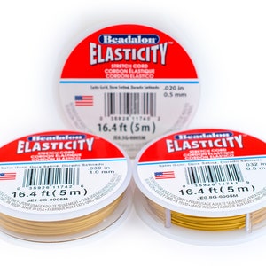 Puede incluir: Tres carretes de cord&oacute;n el&aacute;stico de color dorado, cada uno con una etiqueta blanca que dice "Beadalon ELASTICITY Stretch Cord Cordon Elastique Cordon Elastico". Las etiquetas tambi&eacute;n incluyen el grosor del cord&oacute;n en pulgadas y mil&iacute;metros, y la longitud del cord&oacute;n en pies y metros.