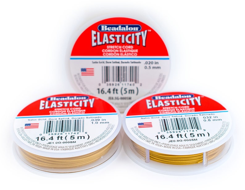 Puede incluir: Tres carretes de cord&oacute;n el&aacute;stico de color dorado, cada uno con una etiqueta blanca que dice "Beadalon ELASTICITY Stretch Cord Cordon Elastique Cordon Elastico". Las etiquetas tambi&eacute;n incluyen el grosor del cord&oacute;n en pulgadas y mil&iacute;metros, y la longitud del cord&oacute;n en pies y metros.
