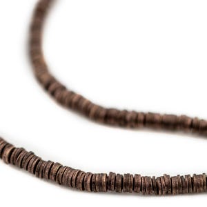 May include: Close-up of a brown beaded necklace against a white background. The necklace is made of small, round, flat beads. The beads are a dark brown color and appear to be made of wood or a similar material. The necklace is in two strands.