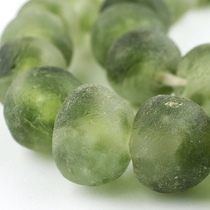 May include: A strand of translucent, olive-green beads. The beads are irregularly shaped and appear to be made of a glass-like material. The beads are strung together, forming a circular shape. The background is white.
