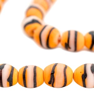 Pode incluir: Fio de contas listradas em laranja e preto. As contas têm formato oval, com uma faixa branca central e listras pretas. As contas são enfiadas em um fio branco.