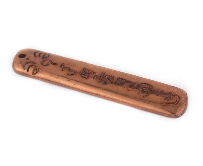 Op de afbeelding: Een koperkleurige hanger met een inscriptie in een onbekende taal. De hanger heeft een gat aan de bovenkant om op te hangen.