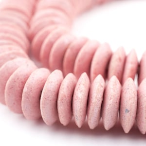 May include: A close-up of a strand of pink, disc-shaped beads. The beads have a slightly rough texture and are arranged in a curved line against a white background. The color is a soft, muted pink.