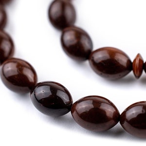 May include: Close-up of a strand of dark brown, oval-shaped beads. The beads have a glossy finish and are strung together, creating a necklace or bracelet. The background is plain white, highlighting the beads' colour and shape.