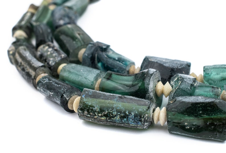 May include: A close-up of a necklace made of dark green and light green glass beads. The beads are cylindrical and have a rough, textured surface. Some of the beads are separated by small, white beads.