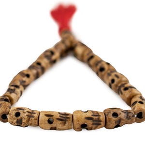 May include: A wooden mala bead necklace with a red tassel. The beads are light brown with dark brown markings and carved details. The necklace is displayed against a white background.