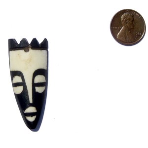 May include: Black and white carved wooden mask with a tribal design. The mask is about the size of a US penny.
