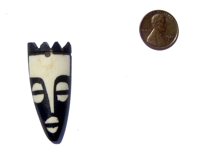 May include: Black and white carved wooden mask with a tribal design. The mask is about the size of a US penny.