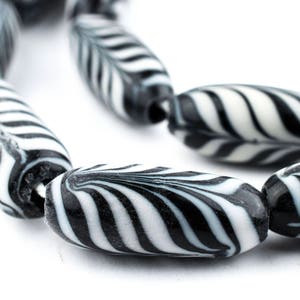 Puede incluir: Primer plano de un collar de cuentas a rayas blancas y negras. Las cuentas oblongas presentan un patrón de líneas negras curvas sobre un fondo blanco. Las cuentas están ensartadas, formando un collar o pulsera.
