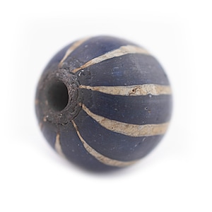 May include: A dark blue, spherical bead with light beige, curved stripes. The bead has a hole through the centre, and the surface appears textured. This decorative bead could be used for jewellery making or other crafts.