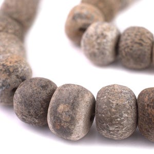 Pode incluir: Close-up de um fio de contas de pedra natural. As contas são ovais e têm uma paleta de cores salpicadas e terrosas de cinza, castanho e bege. As contas são enfiadas, criando uma linha curva.