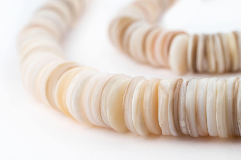 May include: A close-up of a necklace made of flat, round, shell beads. The beads are in shades of cream, beige, and light brown. The necklace is curved, with the beads arranged in a circular pattern against a white background.