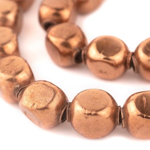 May include: Close-up of a strand of copper-colored beads. The beads are rounded, with a slightly flattened shape, and are strung together. The beads have a metallic sheen and a warm, reddish-brown hue. The background is white.